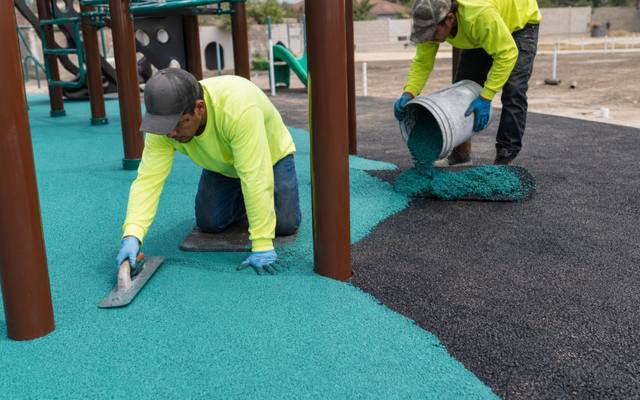 Installer troweling teal rubber surfacing beneath a playground climbing structure.
